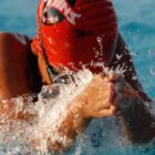 A swimmer in a red swim cap competes in the breaststroke with their hands in front of their face.